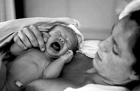 Black and white photograph of a mother holding a new born baby on her chest. The baby's mouth is wide open, crying.